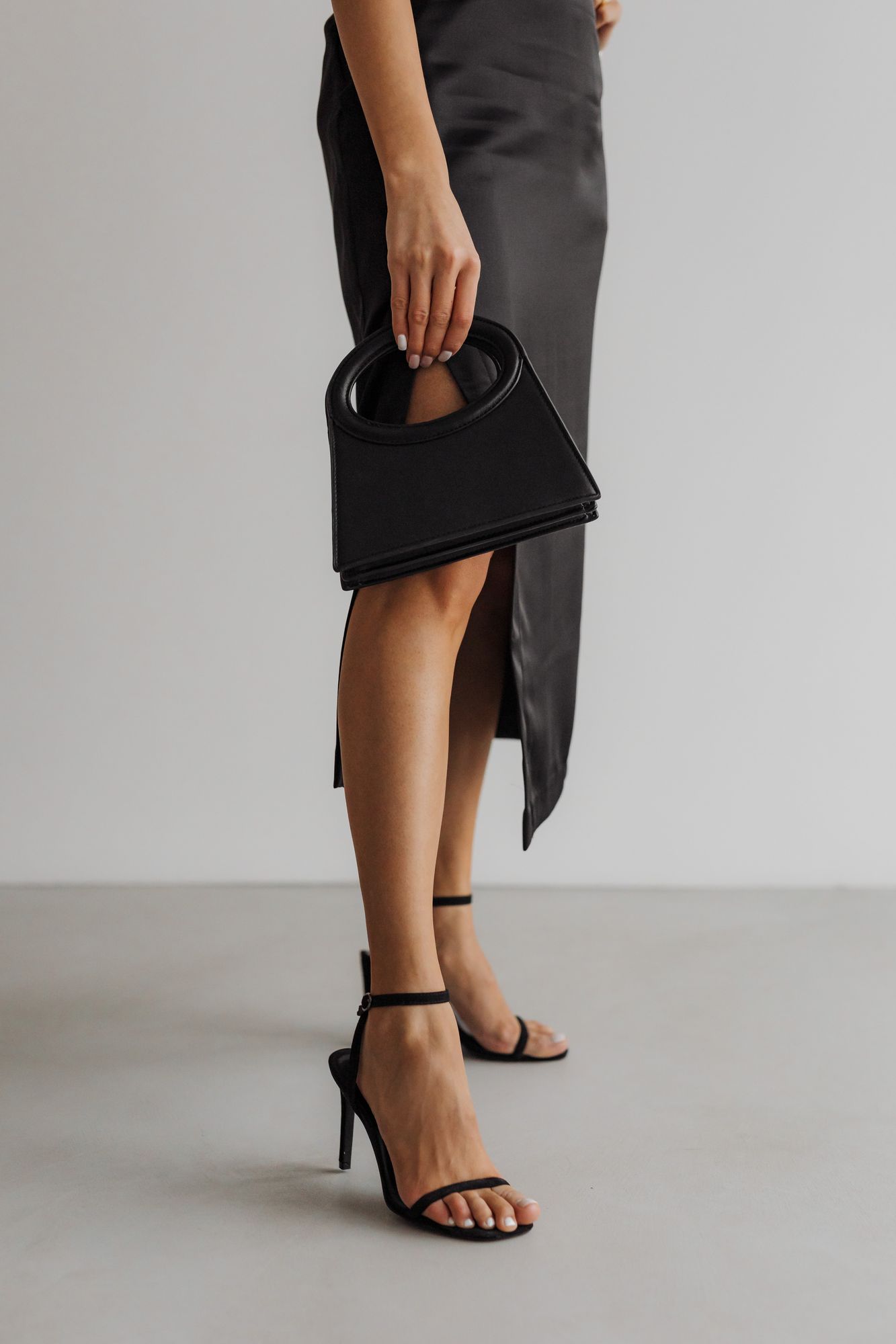 Mulher de vestido de cetim preto segurando uma bolsa estruturada preta, de sandália de tiras